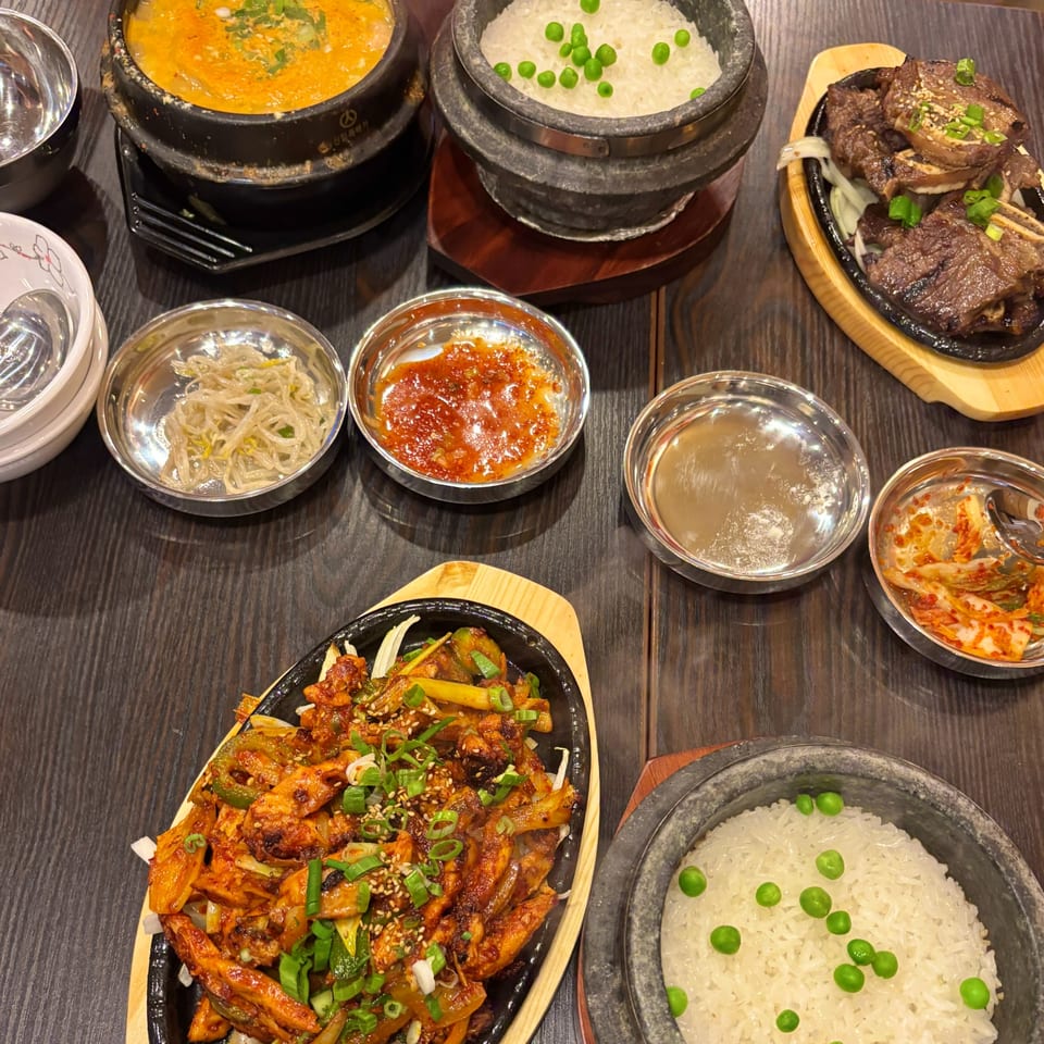 A Korean feast with tofu stew, rice, galbi, spicy chicken, and various banchan on a wooden table.