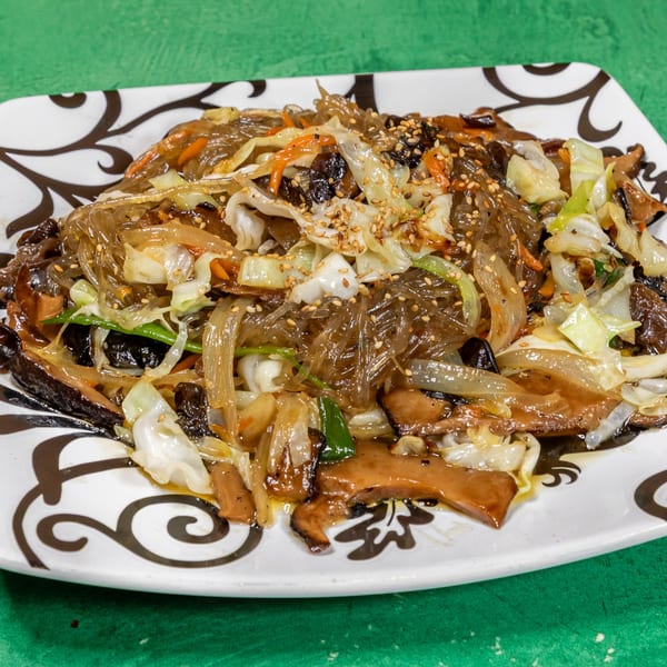 Korean japchae with glass noodles, vegetables, mushrooms, and sesame seeds on a white patterned plate.