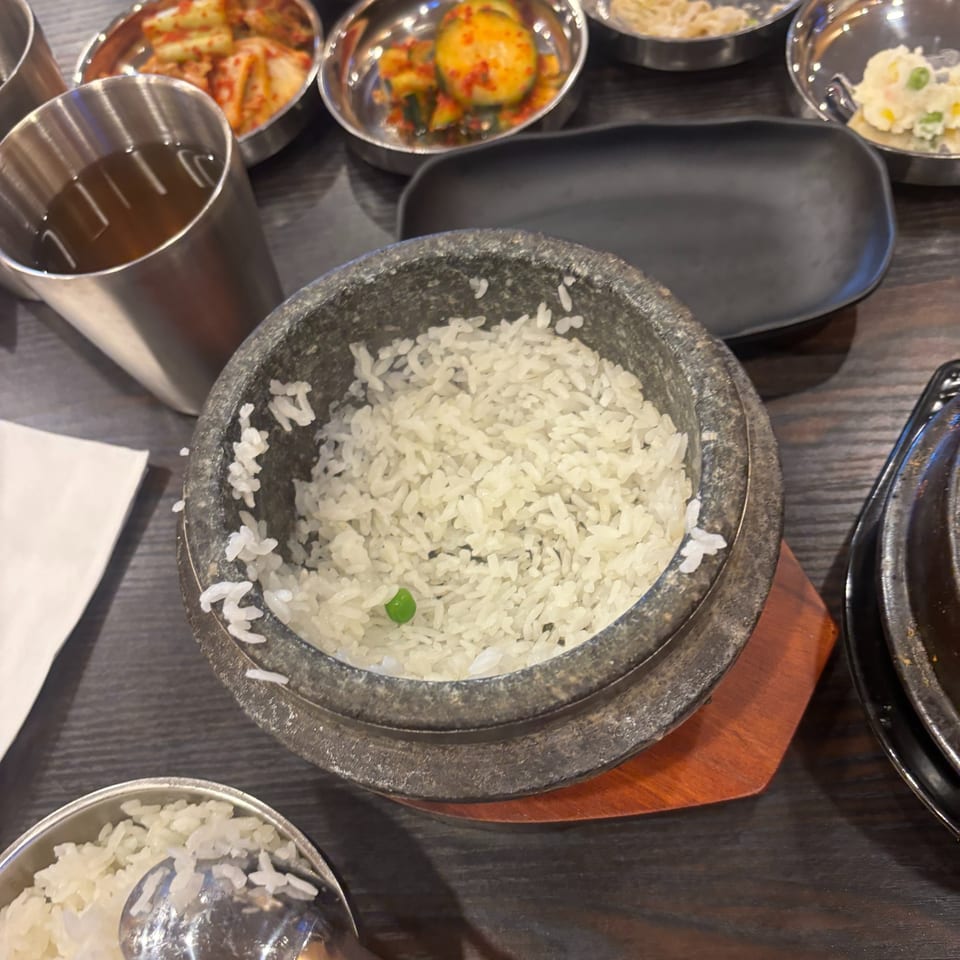 Steaming white rice in a stone pot, with various Korean banchan side dishes and tea.