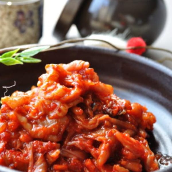 Close-up of a dark bowl filled with red, spicy Korean kimchi, garnished with green leaves.